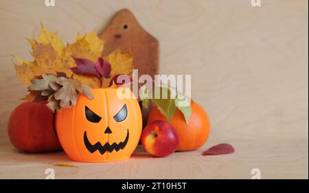 Frohes Halloween. Plastikkübel mit bösem Lächeln und Herbstblättern, hölzernem Hintergrund, Kürbisse, Äpfel und Küchenschneidebrett. Compositi Stockfoto