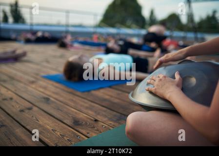 Gruppe junger Menschen, die Yoga-Unterricht üben, liegend in Leichen oder Leichnam im Freien, Shavasana-Übung. Hände mit Trommel im Vordergrund Stockfoto