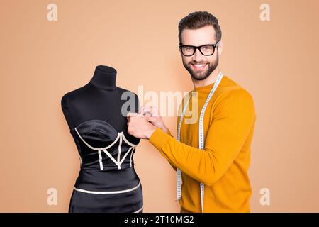 Portrait eines fröhlichen berühmten Schneiders im Pullover mit Zentimeter am Hals, der Messungen von eleganten Kleidern an Schaufensterpuppe mit Blick auf die Kamera durchführt Stockfoto