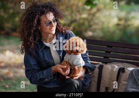 Porträt einer schönen Frau mit lockigen Haaren und einem kleinen Pudel, der lächelt, sitzt draußen im Park. Haustier mit seinem Besitzer in der Natur. Stockfoto