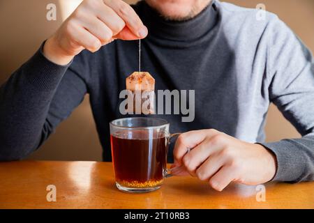 Männlicher Mann, der nassen Teebeutel hält und schwarzen Vitamintee mit Beutel in transparenter Tasse auf dem Tisch brütet. Tee trinken im Café oder zu Hause. Stockfoto