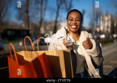 Außenporträt einer glücklichen schwarzen Frau. Sie sitzt nach dem Einkaufen auf der Bank auf der Straße. Stockfoto