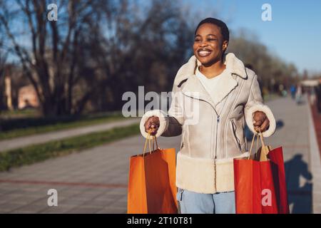 Außenporträt einer glücklichen schwarzen Frau. Sie hält Einkaufstüten in der Hand und läuft auf der Straße herum. Stockfoto