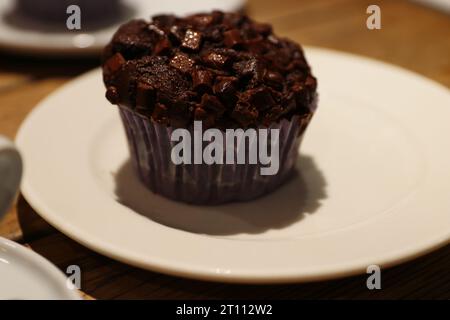 Schokoladenmuffin auf dem weißen Teller. Hausgemachter Cupcake mit Schokoladenchips auf dem Holztisch im Café. Köstlicher Brownie-Kuchen. Gemütliche Atmosphäre. Clos Stockfoto