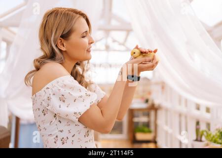 Seitenansicht einer glücklichen blonden Frau, die ein schönes Kleid trägt und mit kleinen gelben Enten posiert, im Sommerhaus mit Pavillon am sonnigen Tag. Stockfoto