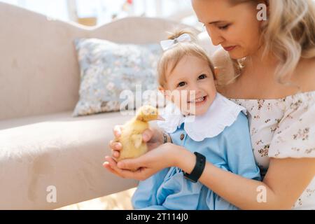 Nahaufnahme Porträt der liebenden jungen Mutter und der liebenswerten Tochter des Kleinkindes im Kleid, die mit kleinen gelben Entlein im Sommerhaus an sonnigen Tagen spielt. Stockfoto