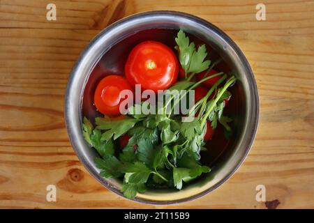 Frisches Gemüse wird in der Metallschale gewaschen, die mit kaltem Wasser gefüllt ist, auf dem Vintage-Schneidebrett aus zerrissenem Holz. Bio-Tomaten, Petersilie, ce Stockfoto