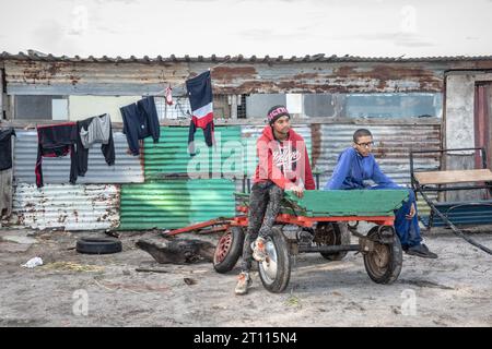 Zwei junge Männer sitzen auf einem Wagen vor einer Wellblechhütte in einer armen Gemeinde in Südafrika Stockfoto