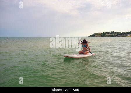 Frau in blauem Bikini sitzt auf einem Sup Board mit einem Paddel im Meer gegen den sonnigen Tag Stockfoto