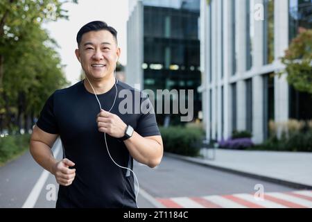Porträt eines jungen asiatischen Mannes, der morgens in Kopfhörern auf einer Stadtstraße läuft und in die Kamera lächelt. Stockfoto