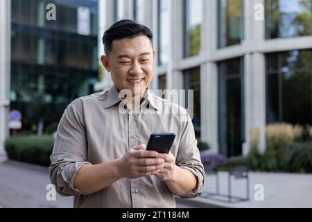 Ein lächelnder junger asiatischer Mann hält ein Handy in der Hand und schreibt Nachrichten, SMS, Anrufe. Lächelnd auf der Straße der Stadt stehen. Stockfoto