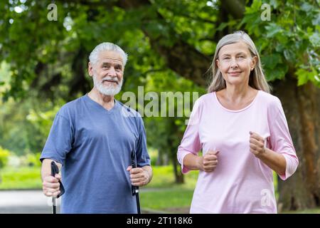 Porträt eines älteren Paares von Mann und Frau, die laufen und Nordic Walking machen. Sie stehen draußen im Park in Sportkleidung und schauen mit einem Lächeln in die Kamera. Stockfoto