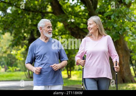 Senior Mann und Frau in Sportbekleidung, die im Park laufen und nordic Walking machen. Sie führen einen gesunden Lebensstil, schauen sich gegenseitig mit einem Lächeln an. Stockfoto