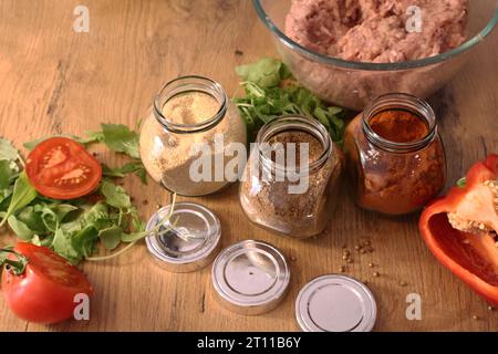 Geräucherter Paprika, getrockneter Knoblauch und Koriander. Gewürzt in den Gläsern auf dem Holztisch. Rote Paprika auf dem Holztisch. Grüne Blätter und Tomaten. Kochen Stockfoto