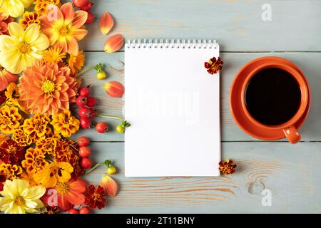 Spiralheft aus Papier mit einer sauberen offenen Seite, einer orangen Tasse schwarzen Kaffees und einer Herbstkomposition und orangen und roten Dahlienblüten, m Stockfoto