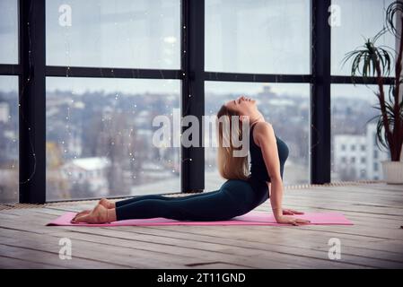 Junge schöne Frau, die Yoga in einem modernen hellen Raum mit großen Fenstern macht. Yoga zu Hause Konzept Stockfoto