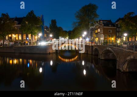 Niederlande. Nachtkanal in Amsterdam. Fassaden typischer Gebäude am Sommerdamm. Beleuchtete Brücke und Verkehrsfluss darauf Stockfoto