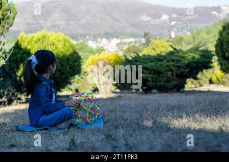 Das kleine Mädchen sitzt am Horizont, in einem Park voller hellgrüner Büsche mit Bergen im Hintergrund. Stockfoto