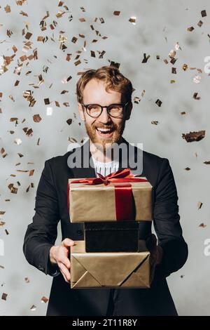 Porträt eines glücklichen jungen Mannes mit einem großen Lächeln, der Geschenke hält, während goldenes Konfetti fallen Stockfoto