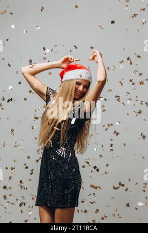 Wunderschönes blondes Weihnachtsmann-Mädchen, das unter glitzerndem Konfetti tanzt. Neujahrsfest-Konzept. Stockfoto