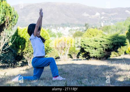 Das kleine lateinische Mädchen praktiziert Yoga im Park, mit einem Hintergrund voller Berge und schöner, hellgrüner Büsche. Stockfoto