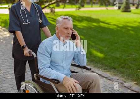 Ein älterer Mann, der einen Patienten im Rollstuhl wiederfindet und am Sommertag mit der Krankenschwester im Park telefoniert Stockfoto
