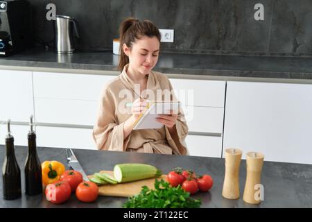 Porträt einer glücklichen jungen Frau schreibt Menü zum Abendessen auf, sitzt in der Küche neben Gemüse, macht Lebensmittellisten zum Einkaufen, posiert im Bademantel Stockfoto