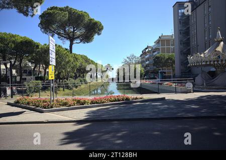 Fröhlich gehen Sie herum und stehen unter zwei Kiefern neben einer Brücke über einen Wasserbach in einer italienischen Stadt an einem sonnigen Tag Stockfoto