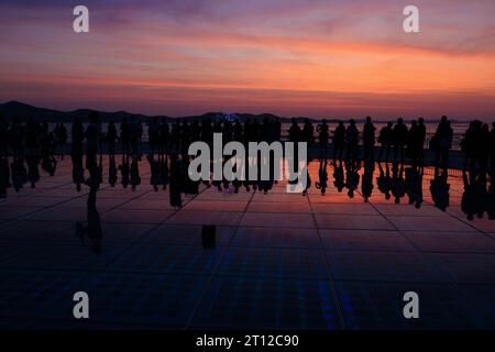 Die Menschenmassen versammeln sich bei Sonnenuntergang um einen Solosaxophonisten, der sich in den Sonnenkollektoren der Begrüßung zur Sonneninstallation bei Sonnenuntergang in zadar kroatien spiegelt Stockfoto