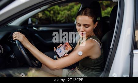 Frau mit Diabetes, die ihren Blutzuckerspiegel überprüft, bevor sie Auto fährt. Diabetikerin, die CGM mit dem Smartphone verbindet, um ihren Blutzuckerspiegel zu überwachen Stockfoto