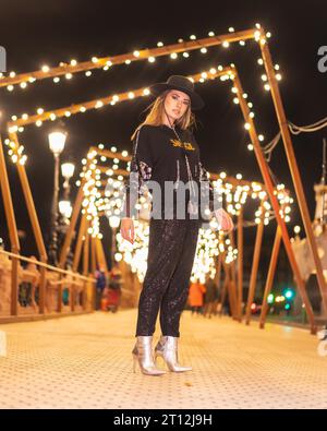 Weihnachtslichter in der Stadt, weißes blondes Mädchen in schwarz gekleidet und mit Pailletten auf einer beleuchteten Brücke, Winterleben Stockfoto