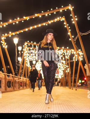 Weihnachtslichter in der Stadt, weißes blondes Mädchen in schwarz gekleidet und mit Pailletten auf einer beleuchteten Brücke, Winterleben Stockfoto