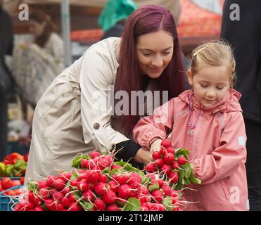 Die junge Frau und ihre kleine Tochter kaufen frisches Gemüse auf einem Straßenmarkt in Prag und pflücken einen Haufen Radieschen von einem großen Stapel. Stockfoto