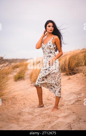 Porträt einer brünetten Kaukasierin, die ihr Haar in einem weißen Blumenkleid streichelt, im Sand am Strand, vertikales Foto Stockfoto