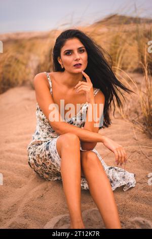 Weißliche Brünette streichelte ihr Haar in einem weißen Blumenkleid, sitzend auf dem Sand am Strand von Cabo de Gata, Nijar. Andalusien, Spanien Stockfoto