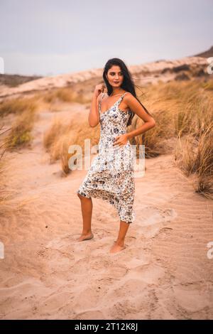 Porträt einer brünetten Kaukasierin, die ihr Haar in einem weißen Kleid streichelt, im Sand am Strand Stockfoto