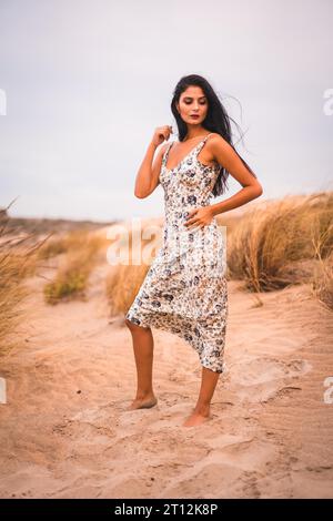 Porträt einer brünetten Kaukasierin, die ihr Haar in einem weißen Kleid streichelt, im Sand am Strand Stockfoto