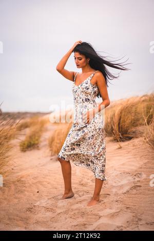 Porträt einer brünetten Kaukasierin, die ihr Haar in einem weißen Blumenkleid streichelt, im Sand am Strand, vertikales Foto Stockfoto