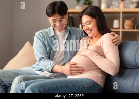 Koreanisches Paar freut sich über die erste Schwangerschaft, sitzt zusammen auf dem Sofa drinnen Stockfoto