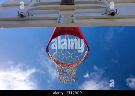 Ein Basketballkorb mit Netz auf einem Platz vor blauem Himmel und flauschigen weißen Wolken bietet die perfekte Umgebung für Outdoor-Sportarten Stockfoto