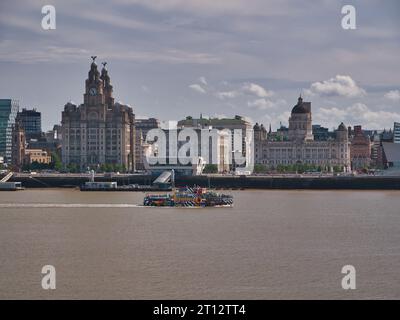 Die River Mersey Ferry Snowdrop passiert die Three Graces an der historischen Hafenpromenade von Liverpool. Das Kunstdesign stammt von Sir Peter Blake. Stockfoto
