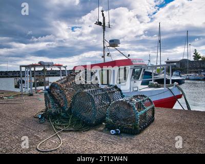 Hummertöpfe (Creels), die an einem Kai neben einem rot-weißen Fischerboot in der Küstenstadt Carrickfergus im County Antrim, Nordirland, gestapelt sind; Stockfoto