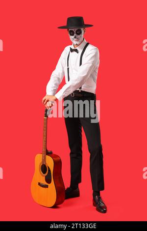 Junger Mann mit bemaltem Schädel und Gitarre auf rotem Hintergrund. Mexikanischer Tag der Toten (El Dia de Muertos) Stockfoto