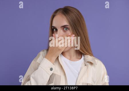 Peinliche Frau, die den Mund mit der Hand auf violettem Hintergrund bedeckt Stockfoto