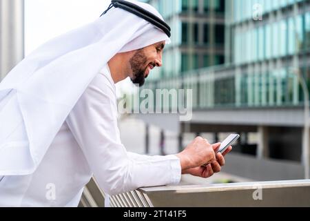 Arabischer Mann aus dem Nahen Osten, der in der Stadt traditionelle emiratische Kandora-Kleidung trägt - arabischer muslimischer Geschäftsmann, der durch das städtische Geschäftszentrum schlendert. Stockfoto