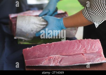 Nahaufnahme von frischem Thunfischbauch. Weibliche Hände mit blauen Handschuhen schneiden große rohe Fische mit einem scharfen Messer im Hintergrund. Vorbereitung für Sashimi. Stockfoto