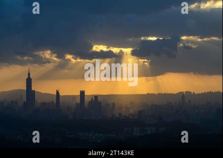 In der Abenddämmerung bricht die Sonne durch die Wolken und scheint auf Taipeh City. Silhouetten von Stadtgebäuden. Orangefarbener Himmel und dunkle Wolken. Stockfoto