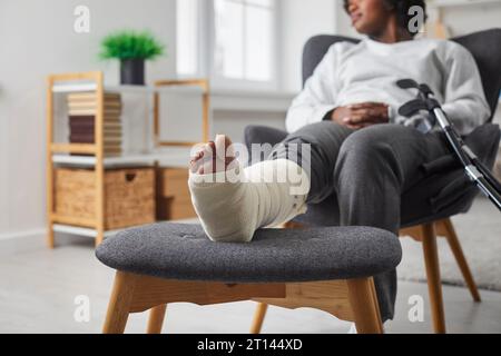 Zugeschnittene Ansicht einer Frau mit Bein in Gipsverband und Krücke im Sessel zu Hause Stockfoto