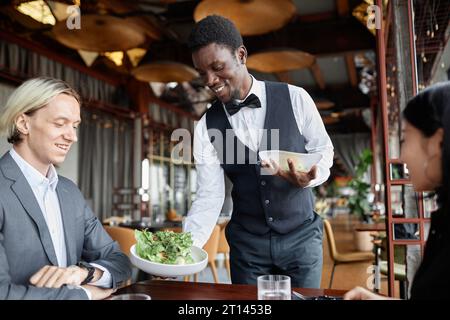 Porträt des jungen Schwarzen Mannes als Kellner, der dem Paar Essen auf Tablett bringt, das im Luxusrestaurant Abendessen genießt Stockfoto