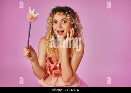 Verblüffte blonde Frau in rosa Kostüm einer Zahnfee, die Zauberstab hält und in die Kamera schaut Stockfoto